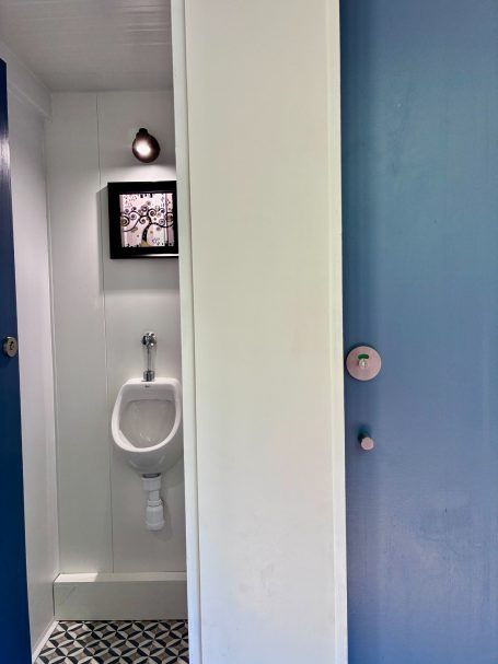 Baño pequeño con un urinario a la izquierda y una puerta azul a la derecha.